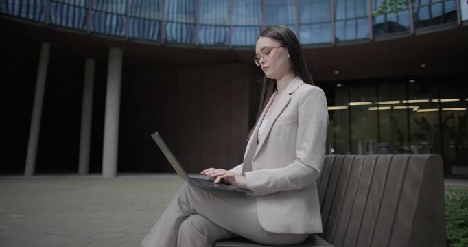 A young Caucasian businesswoman or student or intern in a business suit and glasses in an urban setting among trees and modern buildings sits on a bench and works on a laptop typing a message