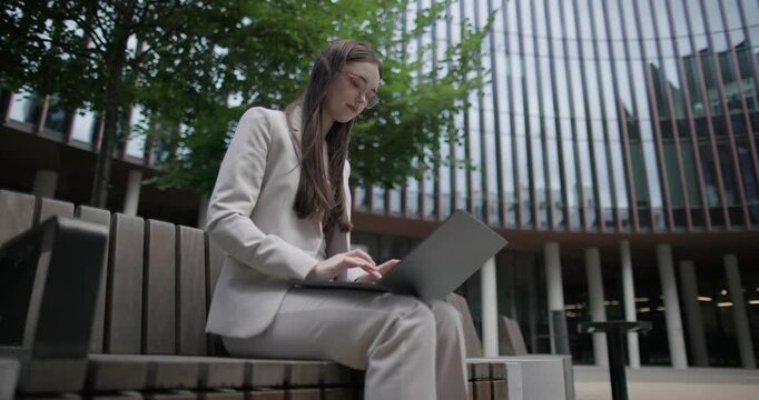 A young Caucasian businesswoman or student or intern in a business suit and glasses in an urban setting among trees and modern buildings sits on a bench and works on a laptop typing a message