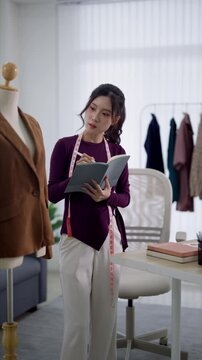 Young Asian female fashion designer reviewing notes in studio surrounded by mannequins with clothing samples and measuring tape around neck