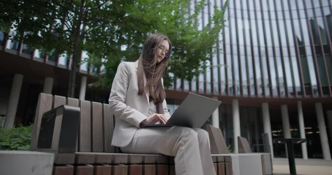 A young Caucasian businesswoman or student or intern in a business suit and glasses in an urban setting among trees and modern buildings sits on a bench and works on a laptop typing a message