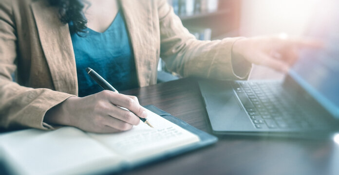 A close-up of a person writing in a notebook while using a laptop. The scene captures a workspace, reflecting productivity and organization in a modern setting.