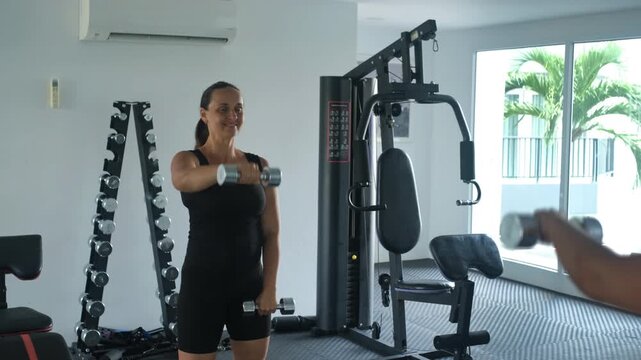 Young woman in black outfit standing in a bright gym lifting dumbbells straight forward in a front raise exercise with weight racks behind her, slow motion. Fitness concept