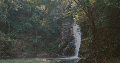 Waterfall cascades down dark rocks into a calm pool surrounded by dense green tropical forest. Natural sunlight filters through trees in a serene wilderness setting. © TravelMedia