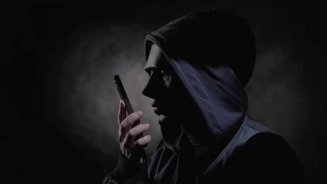 Unrecognizable man in black mask hiding his face speaks on mobile phone, threatening call