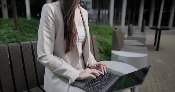 A young Caucasian businesswoman or student or intern in a business suit and glasses in an urban setting among trees and modern buildings sits on a bench and works on a laptop typing a message