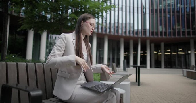 A young Caucasian businesswoman or student or intern in a business suit and glasses in an urban setting among trees and modern buildings sits on a bench and conducts a video call on a laptop