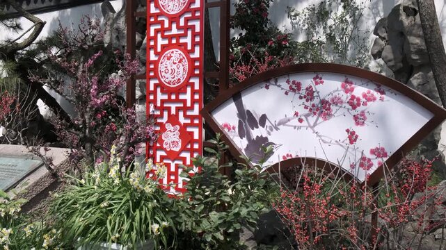 Ornate red paper-cut patterns and a large fan-shaped illustration of blossoms decorate a traditional Chinese garden landscape filled with blooming flowers and scholar rocks.
