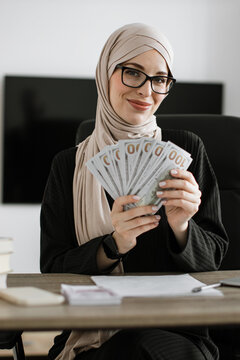 A smiling woman wearing a hijab and glasses holds a fan of US dollar bills while sitting at a desk with papers and a pen