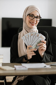 A smiling woman wearing a hijab and glasses holds a fan of US dollar bills while sitting at a desk