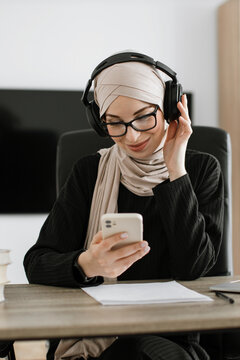 A woman wearing a hijab and headphones is looking at her smartphone while sitting at a desk with papers