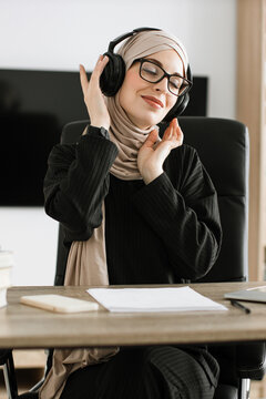 A woman wearing a hijab and glasses enjoys music through headphones while sitting at her desk