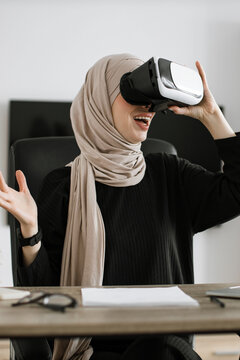 A smiling woman wearing a hijab and VR headset is seated at a desk, experiencing virtual reality