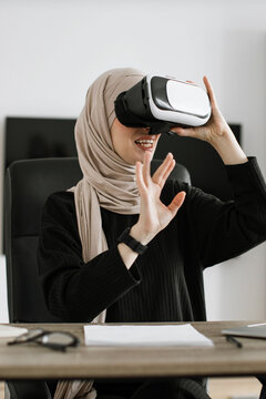 A woman wearing a hijab and a virtual reality headset is actively engaged in an immersive digital experience while seated at a desk