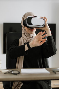 A woman wearing a hijab and a virtual reality headset is seated at a desk, reaching out with her hands as if interacting with a digital environment
