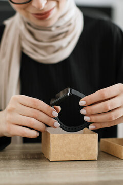 A woman wearing a hijab and glasses carefully removes a new fitness tracker from its packaging box