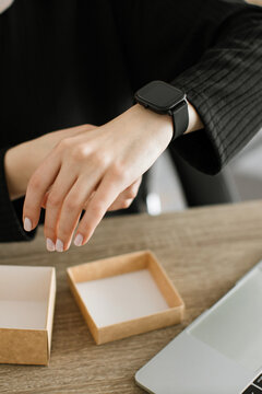A woman's hand wearing a black smartwatch is shown on a wooden desk next to an open box and a laptop