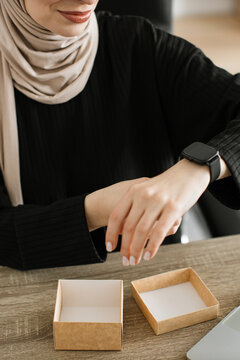 A woman wearing a hijab and a black top checks her smartwatch while sitting at a wooden desk with an open box and a laptop