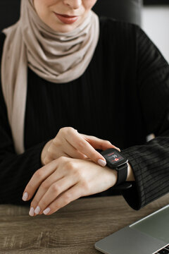 A woman wearing a hijab and black top checks her smartwatch displaying a heart rate reading while working at a desk
