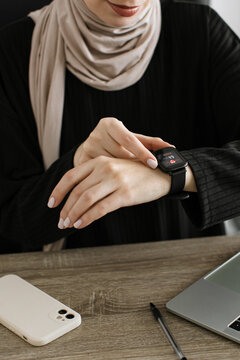A woman wearing a hijab and black top interacts with her smartwatch displaying a heart rate reading while seated at a wooden desk with a laptop and phone