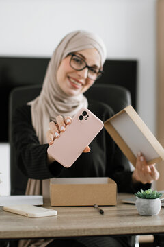 A smiling woman wearing a hijab and glasses unboxes a new pink smartphone from its packaging
