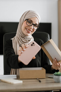 A smiling woman wearing a hijab and glasses holds up a new pink smartphone she just unboxed from a cardboard package