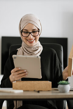 A smiling woman wearing glasses and a hijab is unboxing a package while looking at a tablet device in an office setting