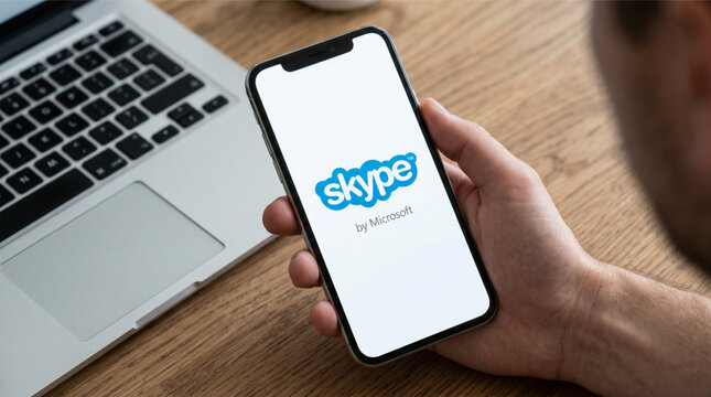 Man holding a smartphone with the Skype by Microsoft app logo on the screen over a wooden desk with a laptop. Communication technology, video calling, and remote business workspace concept mockup.