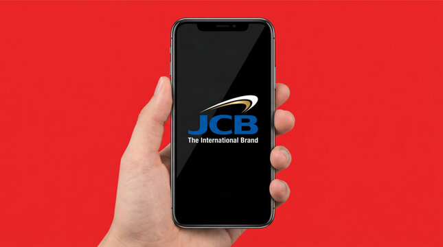 Hand holding a smartphone with the JCB logo on the screen, known as The International Brand. Isolated on a vibrant red background. Mobile payment, credit card application, finance, digital wallet.