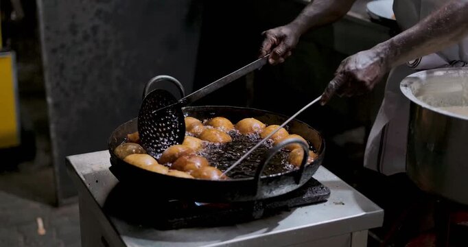 Cinematic close-up of a street food vendor deep-frying traditional Indian snacks, known as Bonda or Pakoda, in boiling oil inside a large iron kadhai (wok). The golden-brown fritters sizzle under the 