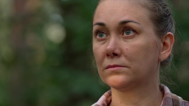 Close-up portrait of a distressed and concerned woman with her hair pulled back, looking up with a worried and anxious expression while standing outdoors in a natural green environment