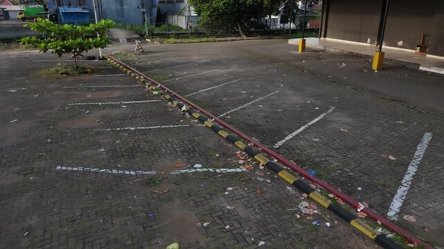 An empty urban parking lot with faded lines, trash scattered about, and an industrial area in the background.