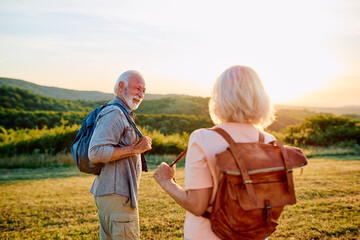 Naklejka na ściany i meble Happy senior couple hiking on a grassy hill, carrying backpacks, and enjoying a beautiful sunset in nature