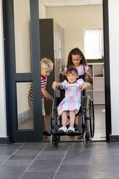Three school-age children pushing manual wheelchair through doorway at classroom with tiled floor