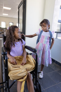 Diverse schoolgirls chatting at class door, one sitting in wheelchair, yellow and blue backpacks
