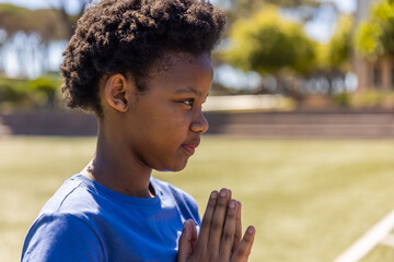 Fototapeta premium African American schoolboy standing on field near white line facing right, wearing blue tee praying