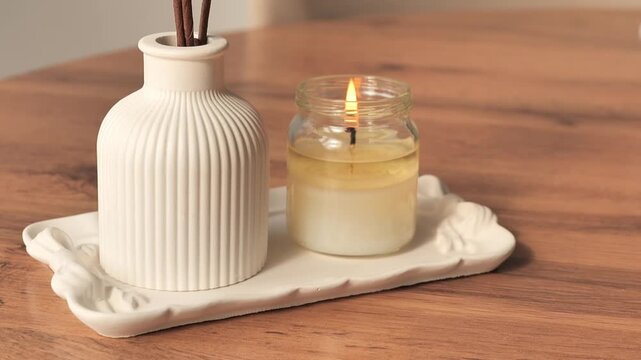 Close-up video of a burning scented candle and reed diffuser placed on a ceramic tray in a cozy home interior. Soft warm lighting creates a relaxing atmosphere associated with aromatherapy, wellness