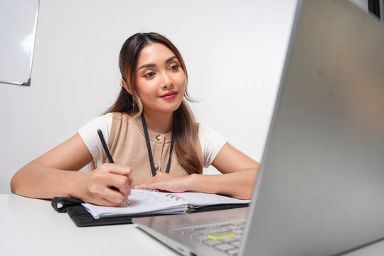 Attractive young Asian woman is working looking at her laptop and writing a report on book.