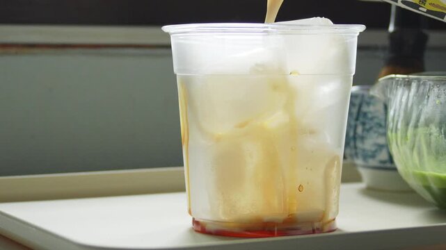 4K Close up of pouring creamy brown liquid over ice cubes in clear plastic glass on white tray, Step of preparing refreshing iced beverage.