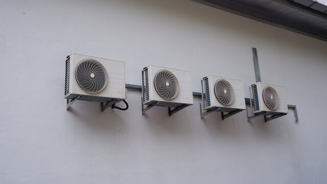 a landscape low-angle shot of four unbranded outdoor air conditioner compressor units mounted on a plain white concrete wall in bright daylight.