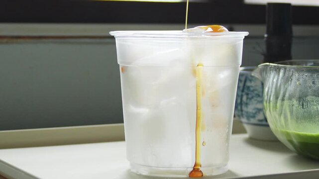 4K Close up of pouring creamy brown liquid over ice cubes in clear plastic glass on white tray, Step of preparing refreshing iced beverage.