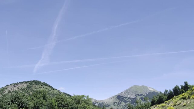 Paesaggio montano verde e soleggiato, con due rilievi ricoperti di vegetazione e cielo limpido attraversato da scie aeree