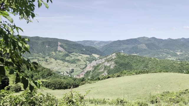 Ampio paesaggio collinare e montano, con prati verdi, vallate luminose e catene montuose che sfumano all&rsquo;orizzonte sotto un cielo sereno