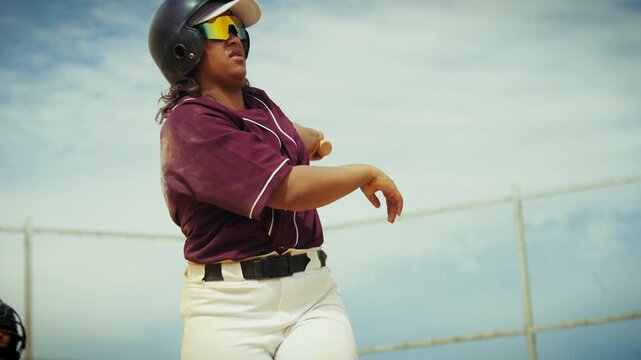 A female athlete in a baseball uniform swings powerfully, making solid contact with the ball. She watches the flight, showing dedication and focus.
