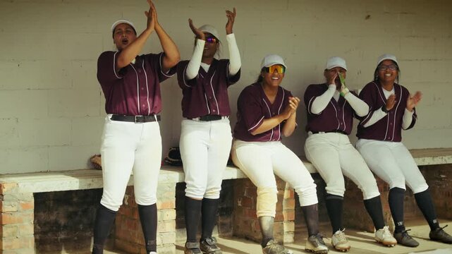 Enthusiastic female baseball players in uniform cheer and clap from the dugout, showcasing team spirit and support during a game.
