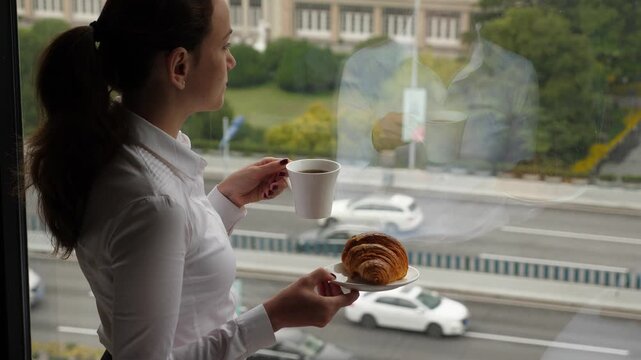 Thoughtful woman stay with cup of coffee and croissant against window, looking outside. City traffic seen down on the road. Lady thinking and stare to distance, fixed at one place