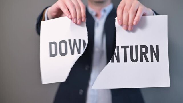 A male adult in formal attire tears a sheet of paper with the word downturn printed on it. This action occurs in a neutral setting, conveying the concept of confronting economic obstacles