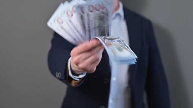 A person offers a bundle of cash with their right hand while standing against a plain background