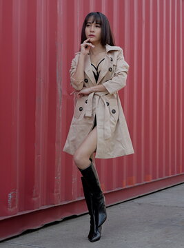 Young woman in a beige trench coat and black lingerie stands with her hand on her chin near a red shipping container background