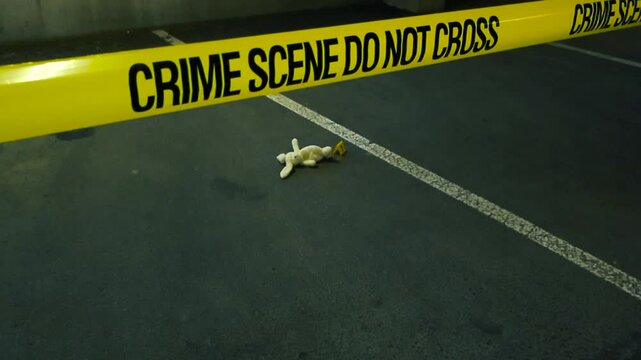Abandoned stuffed bunny toy lying on the ground behind yellow caution tape marking a crime scene in an underground parking garage, representing a child abduction or kidnapping