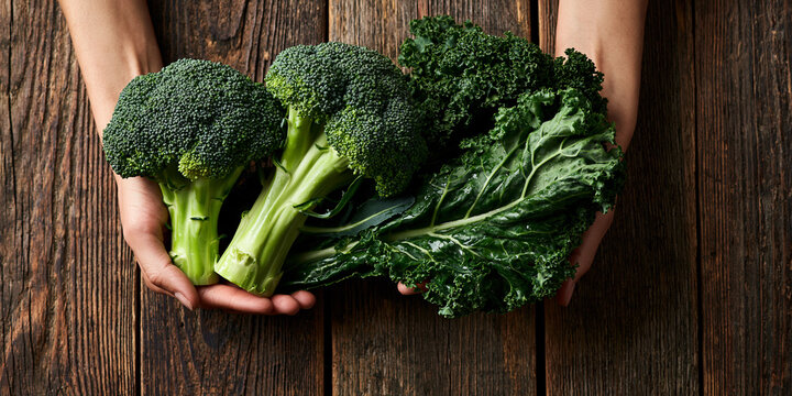 Fresh broccoli and kale held in hands over a rustic wooden table